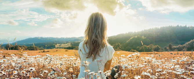 A woman sitting in a field of white flowers, looking out towards distant hills beneath a bright, cloudy sky.